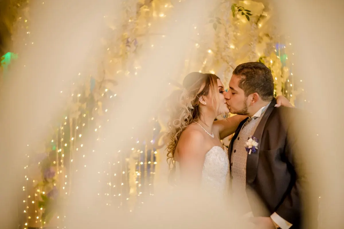 A bride and groom share a romantic kiss surrounded by enchanting lights, capturing the essence…