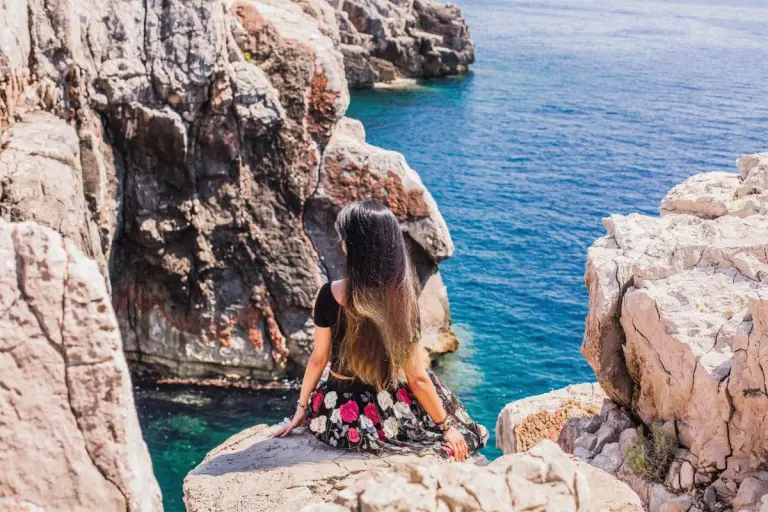 A woman sits on a rocky cliff, gazing at the clear blue Adriatic Sea in Croatia. - Photo by…