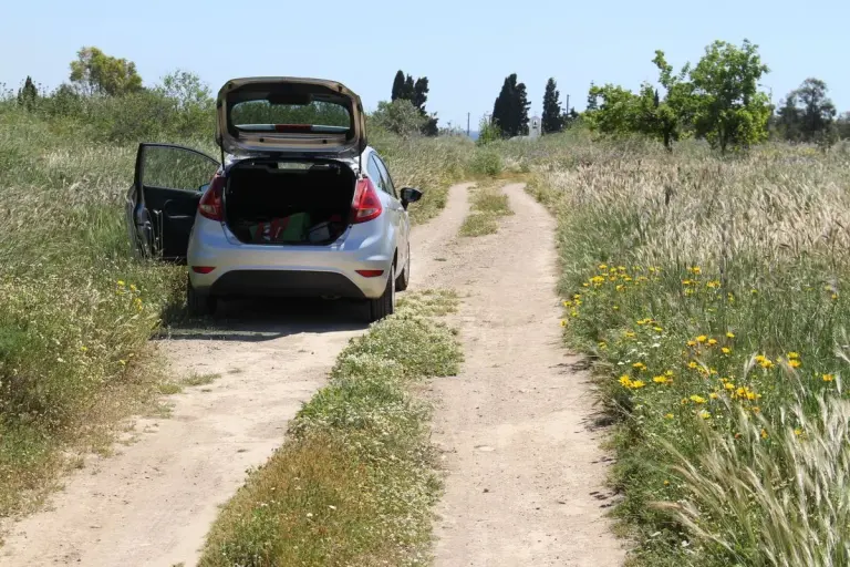 A car parked on a countryside dirt road surrounded by a grassy field under clear skies. - Photo…