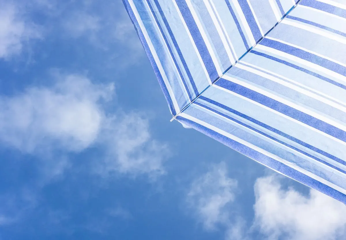 A striped beach umbrella set against a vibrant blue sky with fluffy clouds, conveying summer…