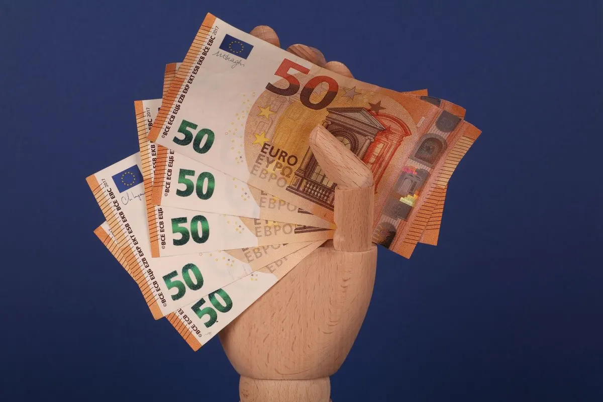 A wooden mannequin hand grasping fifty euro banknotes with a blue backdrop. - Photo by Ann H on…