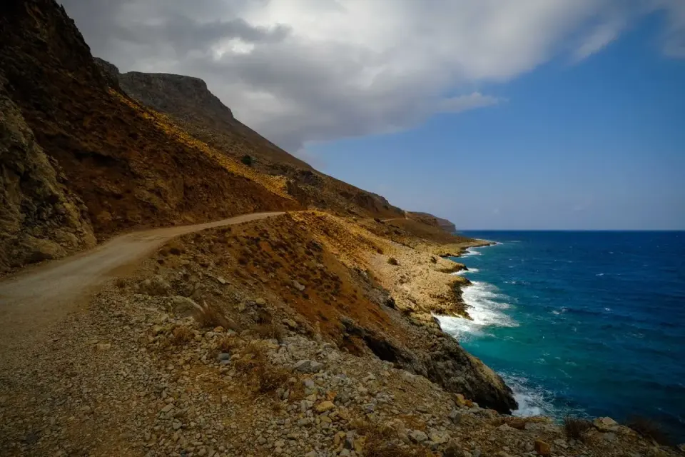 Rustic coastal road winding along a rocky cliff with ocean views under a blue sky. - Photo by…