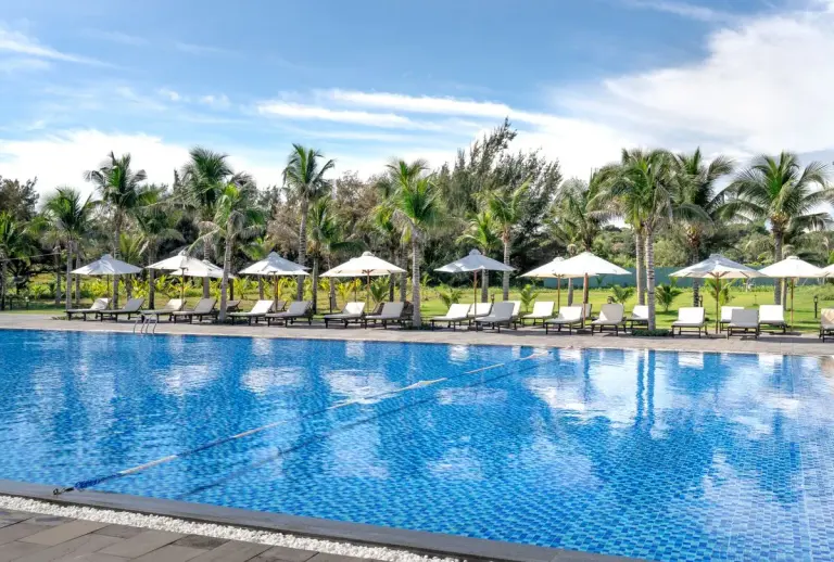 Relaxing poolside scene with umbrellas, palm trees, and loungers under a clear blue sky. - Photo…