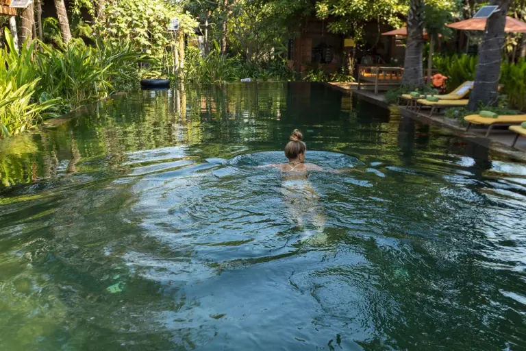 A woman enjoys a swim in a serene tropical resort pool surrounded by lush greenery. - Photo by…
