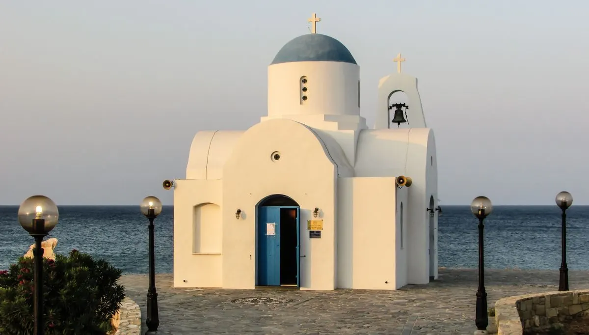 A traditional white chapel with blue dome and cross by the seashore, ideal for travel and…