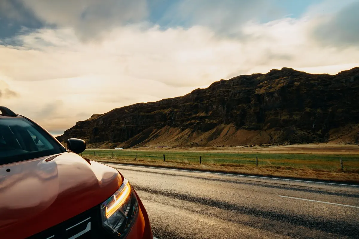 A red SUV travels along a rural road with a picturesque mountain landscape under a bright sky. -…