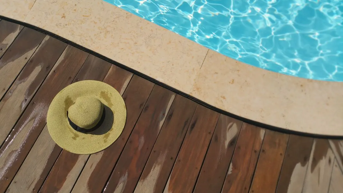 A straw hat rests on a wooden deck beside a sparkling blue swimming pool, perfect for summer…