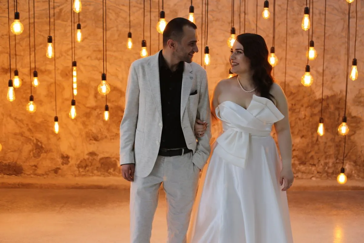 Couple in elegant attire, surrounded by warm hanging lights, embracing joy. - Photo by İSMAİL…