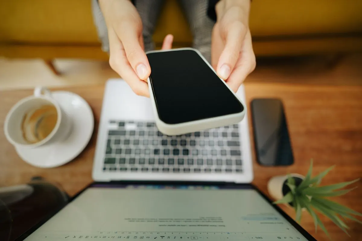 Hands holding smartphone over laptop in cozy home office setup with coffee and plant. - Photo by…