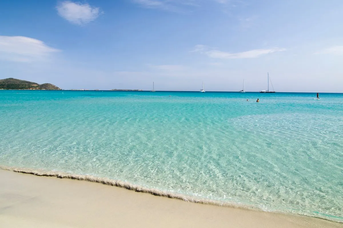 Tranquil beach scene with pristine turquoise waters and distant sailboats under a sunny sky. -…