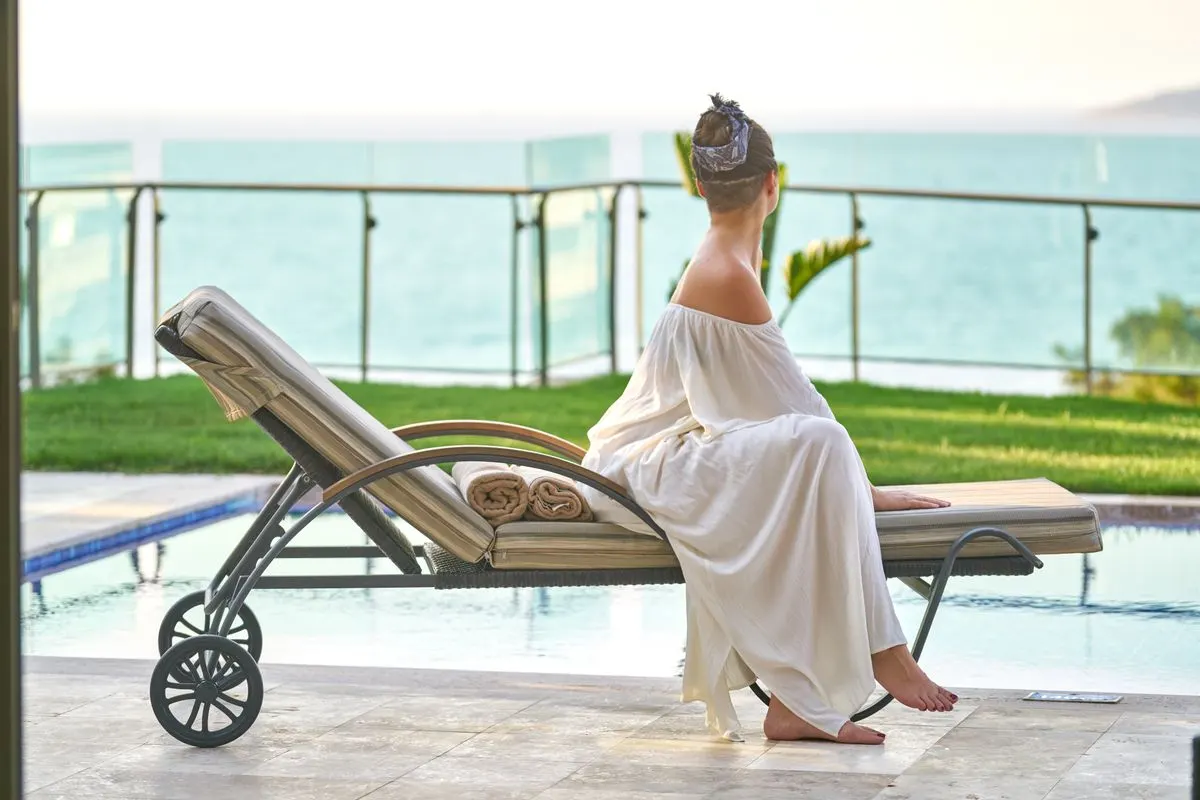 A woman in a white dress relaxes by the poolside with an ocean view. - Photo by Engin Akyurt on…