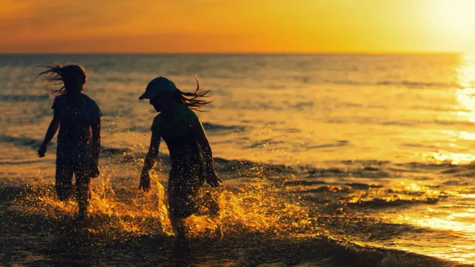 Two children joyfully splash in the water at a beach during sunset, capturing carefree summer…