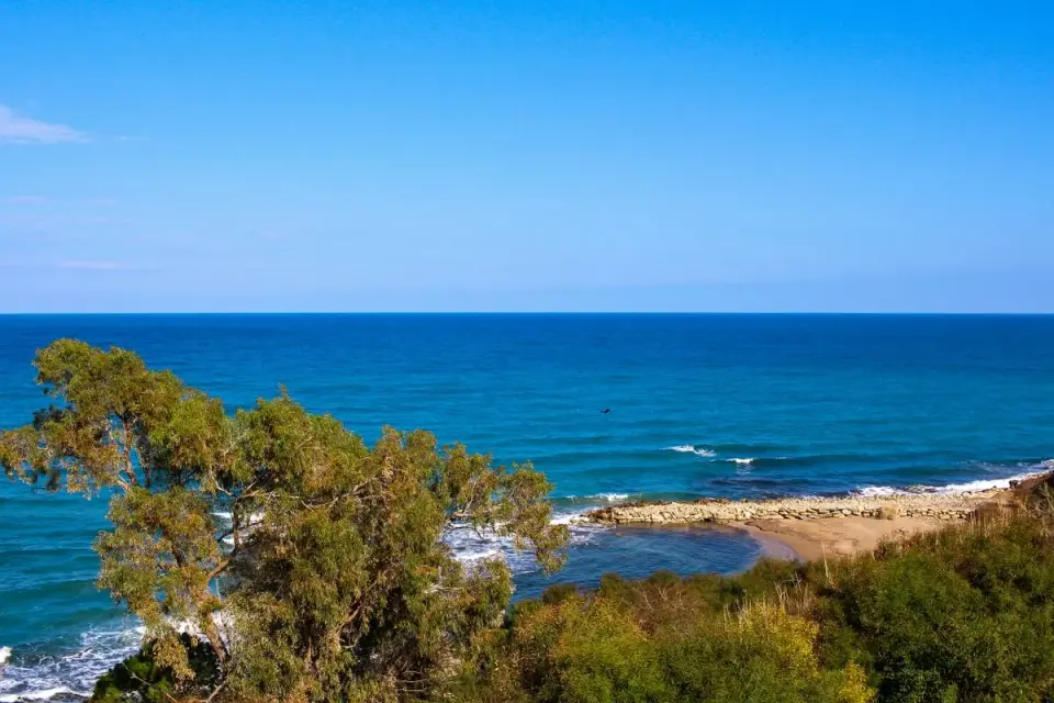 A breathtaking view of a blue ocean with a rocky shoreline and lush greenery under a clear sky.…