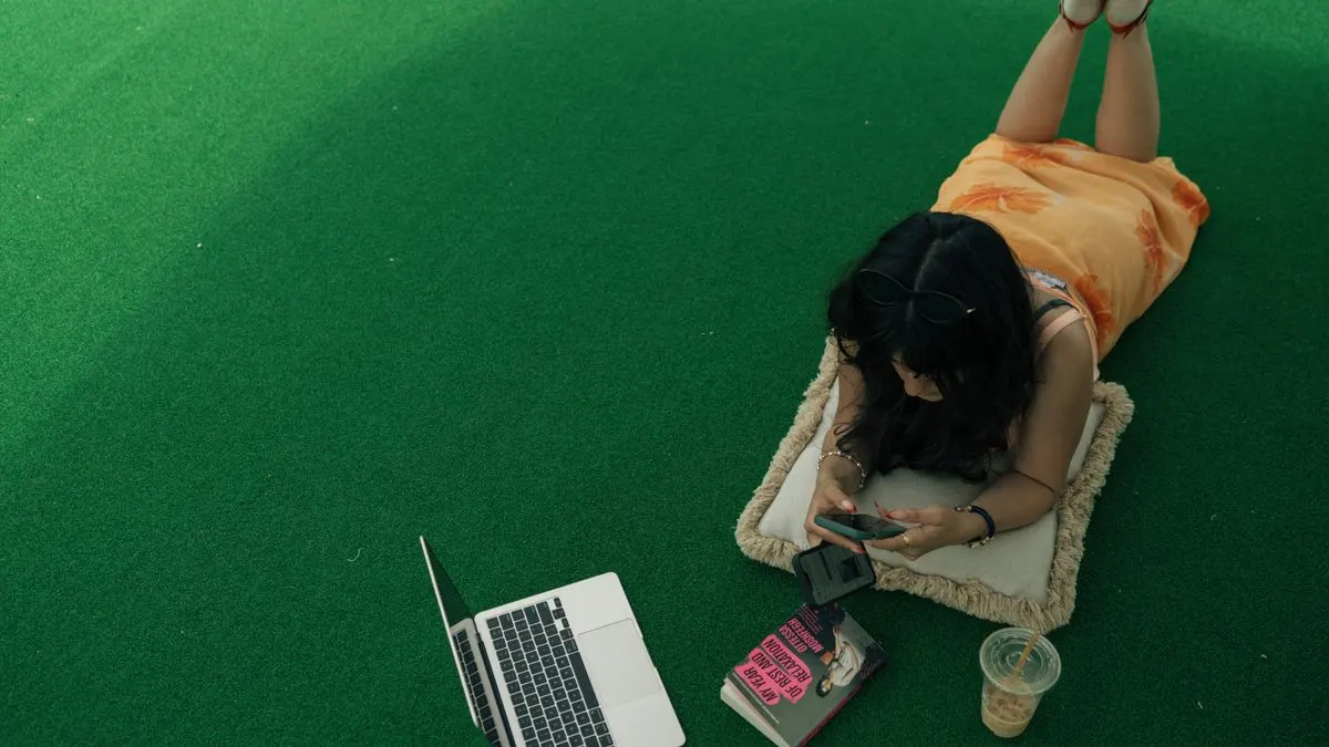 Woman lying on a cushion outdoors using phone, with laptop and book nearby. - Photo by Sami…