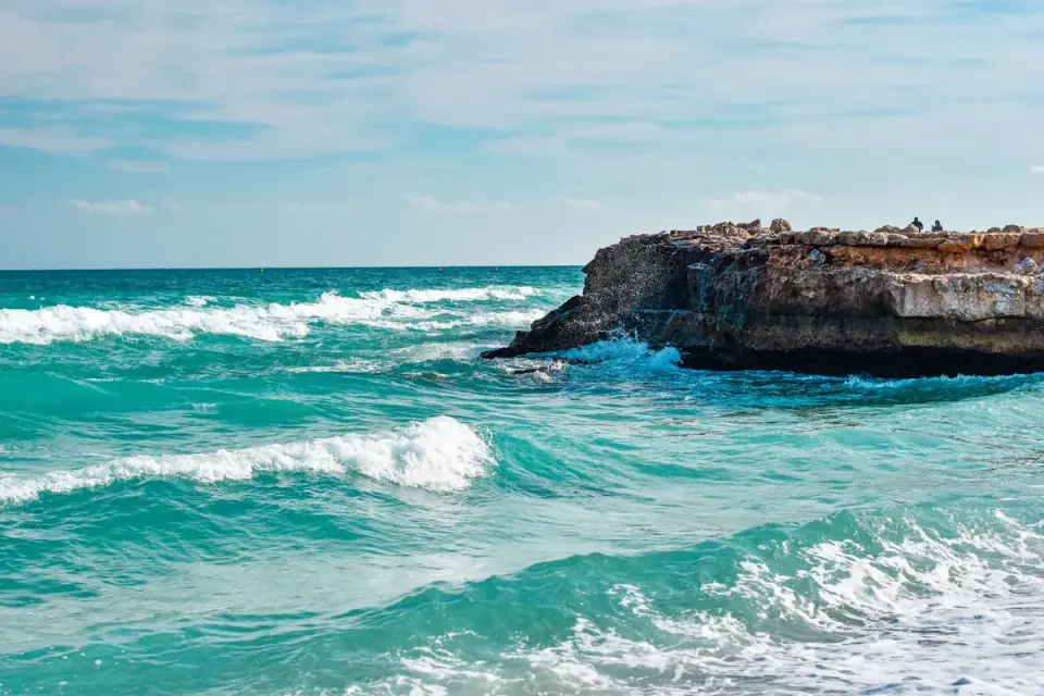 Vibrant ocean waves crashing against a rocky cliff under clear blue skies, showcasing nature's…