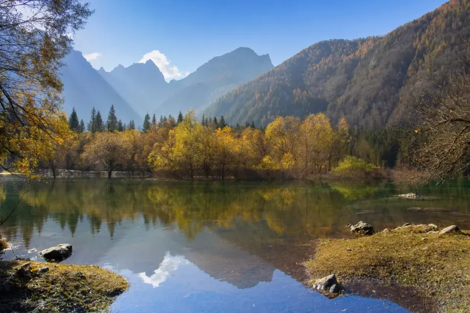 A tranquil alpine lake surrounded by autumn-colored trees and majestic mountains. - Photo by…