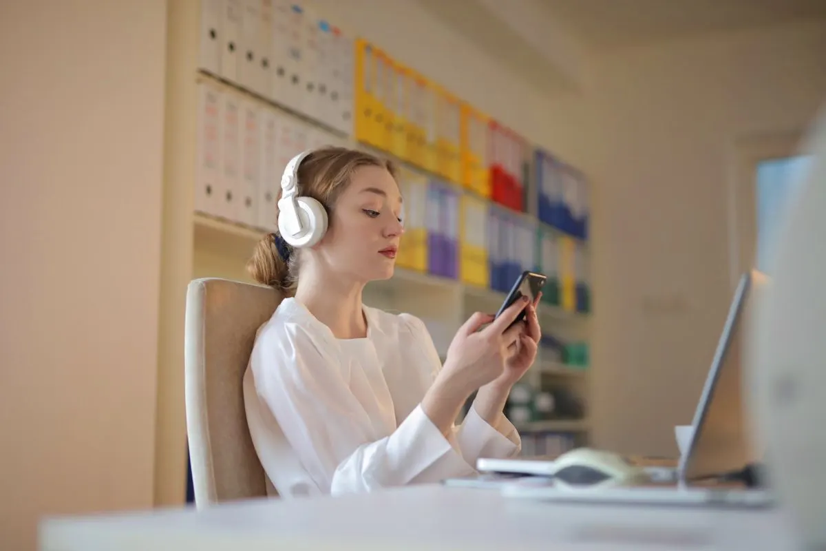 Young woman in white shirt using smartphone and headphones at her office desk. - Photo by Andrea…