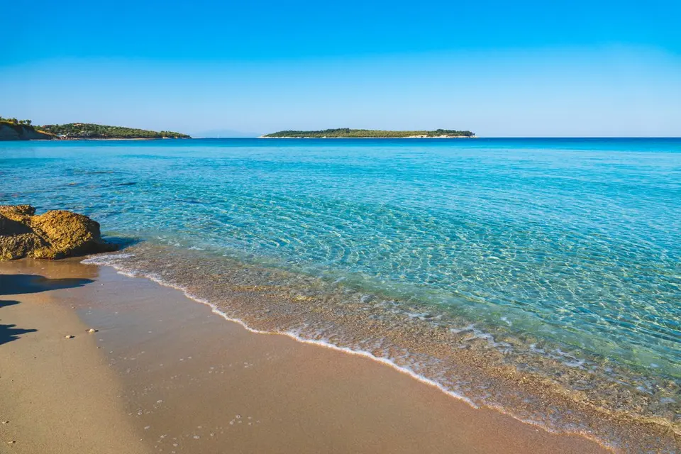 Peaceful sandy beach with crystal clear water and distant islands under a bright blue sky. -…