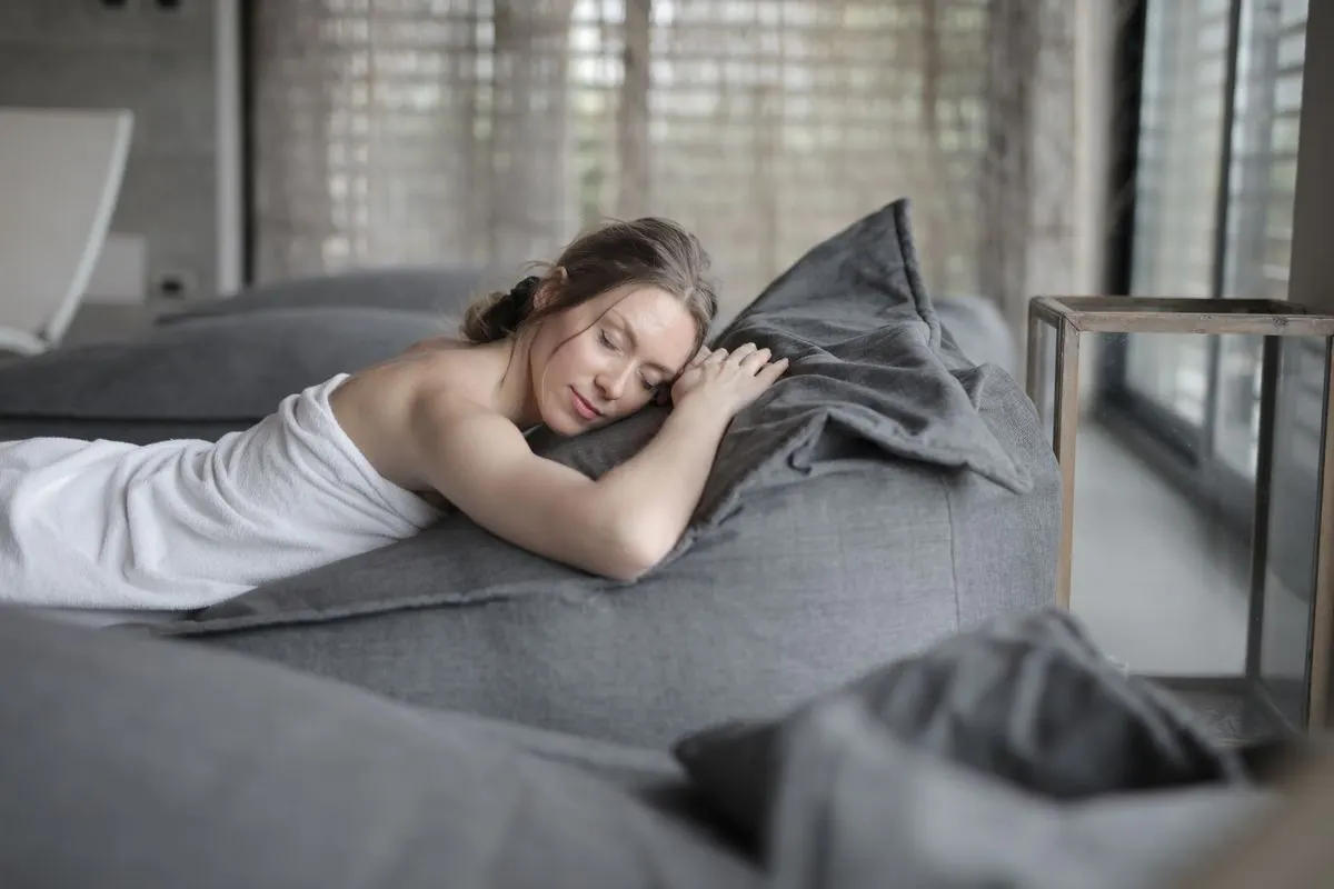 Woman peacefully relaxing at a spa, wrapped in a towel on a comfortable bed, enjoying serene…
