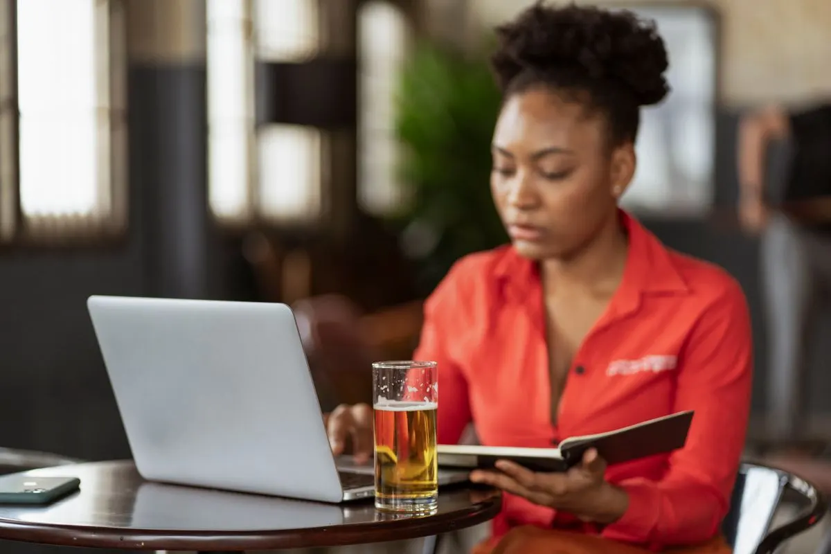 Woman sitting at a table with a laptop and book, having a drink. Indoor setting. - Photo by…