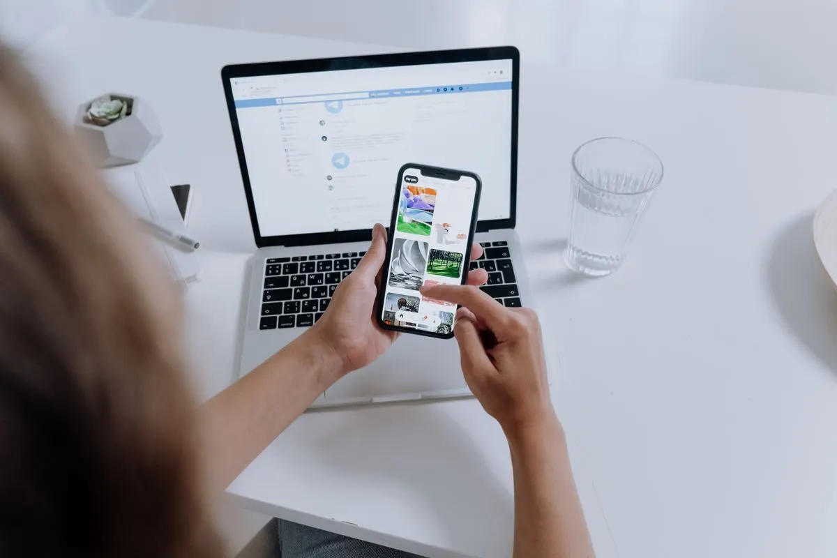 Person using both a smartphone and laptop, multitasking at a modern desk with office plants. -…