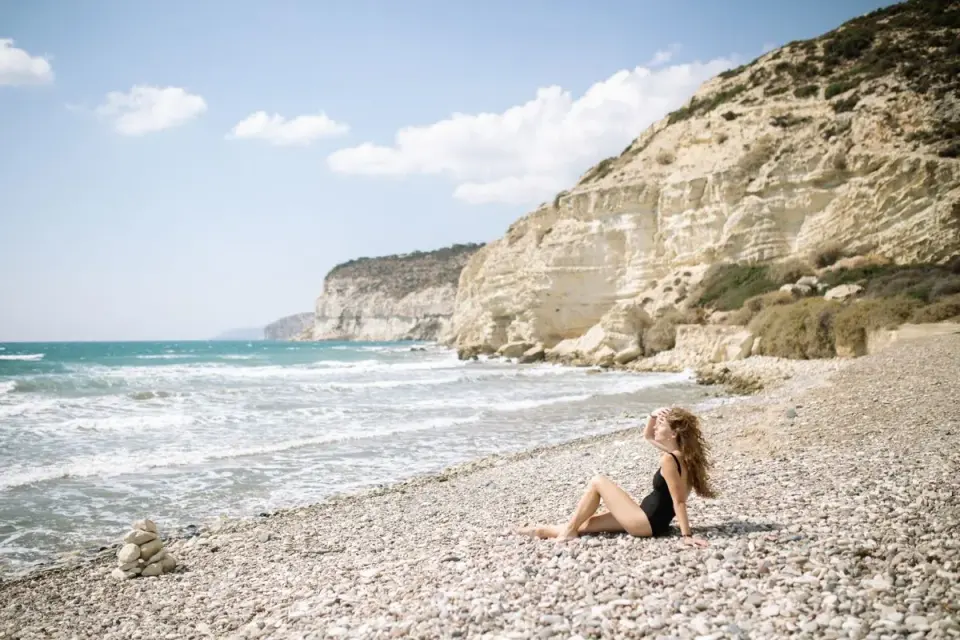 A woman in a black swimsuit relaxes on a pebble beach by an ocean under clear skies. - Photo by…