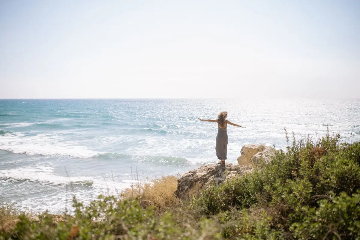 A woman stands on a rocky cliff with open arms overlooking the ocean, radiating freedom and…