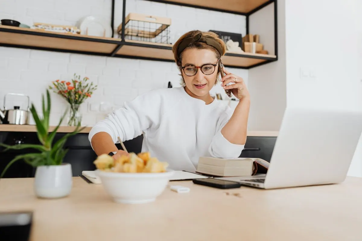 Adult woman in a white sweatshirt working from home office with laptop and phone, in a stylish…