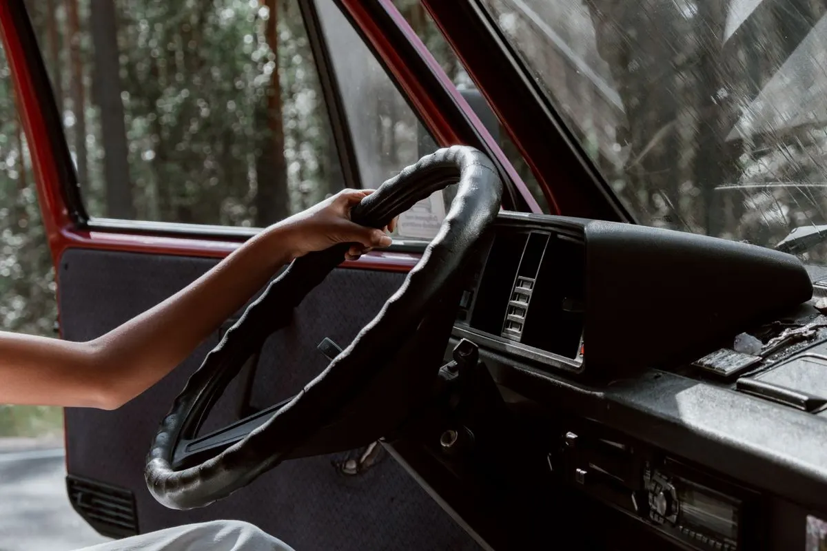 A close-up of a hand gripping a steering wheel in a vintage vehicle, surrounded by forest. -…