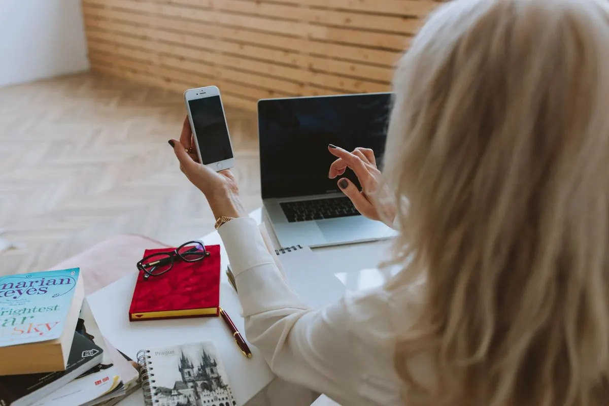 Businesswoman multitasking with a smartphone and laptop, working indoors at a wooden desk. -…