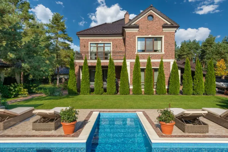 Facade of brick dwell villa with green bushes on lawn and sunbeds near swimming pool - Photo by…