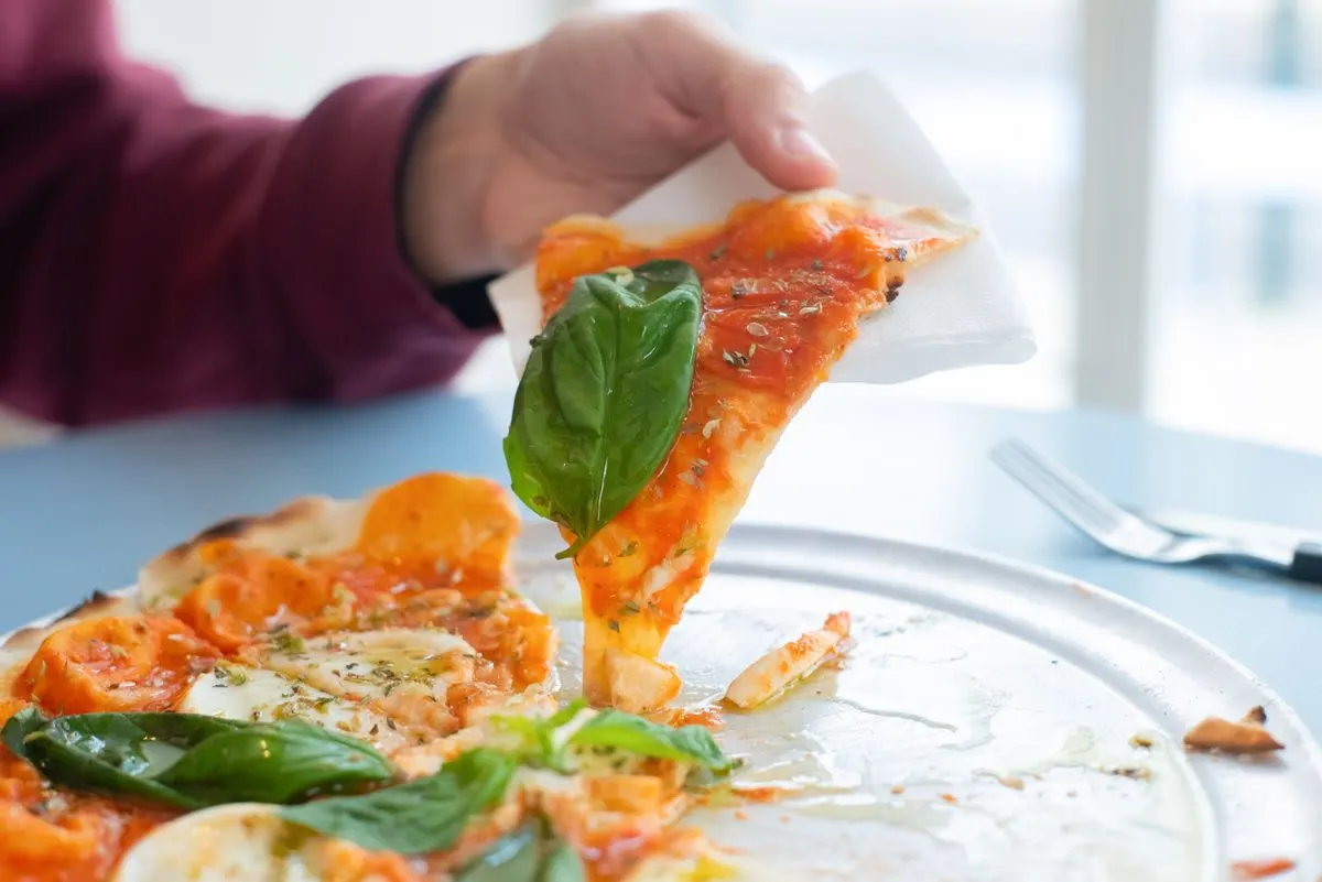 Close-up of a hand holding a slice of delicious Margherita pizza with basil topping on a sunny…