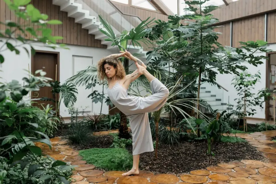 A woman performs a yoga pose amidst lush greenery in a tranquil indoor garden setting. - Photo…