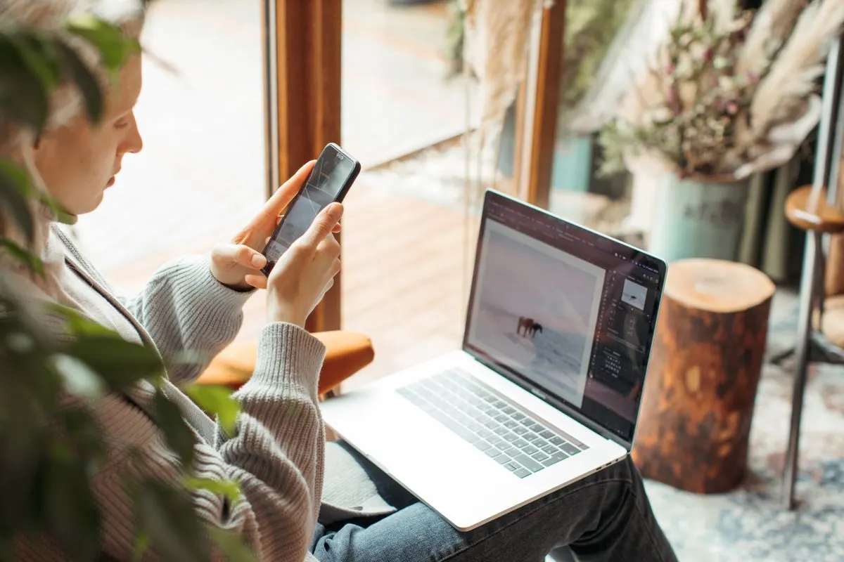 A woman using her smartphone and laptop for remote work indoors, capturing a cozy workspace. -…