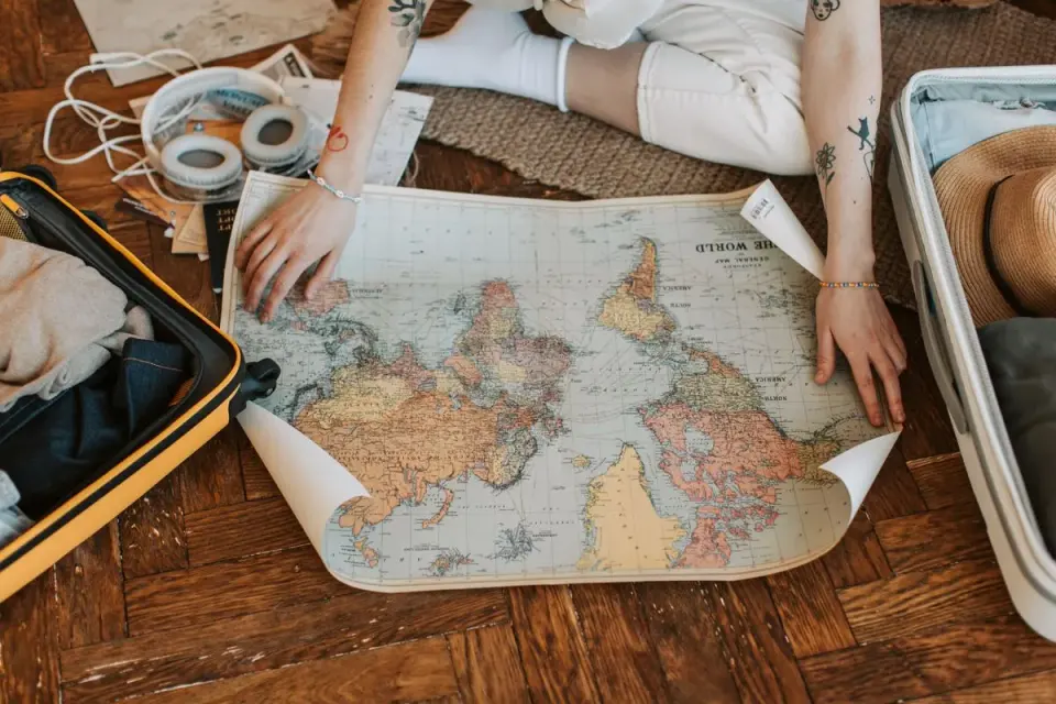 A traveler plans a journey, sitting on the floor with a world map and suitcase filled with…