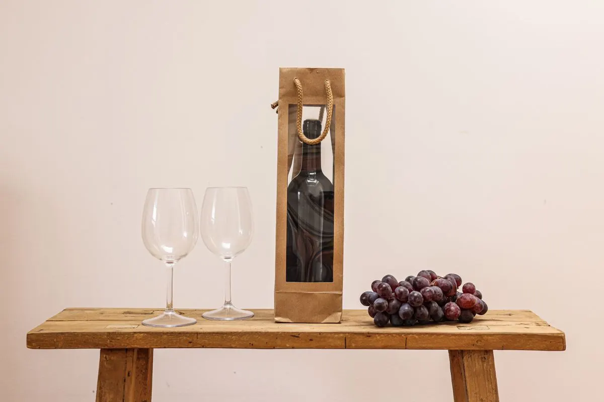 A classic still life of a wine bottle in a bag, wine glasses, and grapes on a wooden table. -…