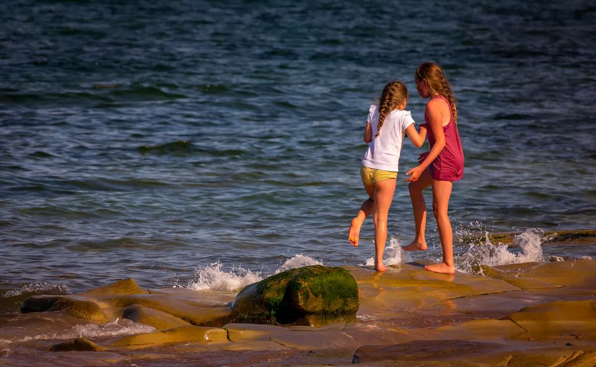 Two girls joyfully playing on a rocky shore by the sea during a sunny day. - Photo by Ray…