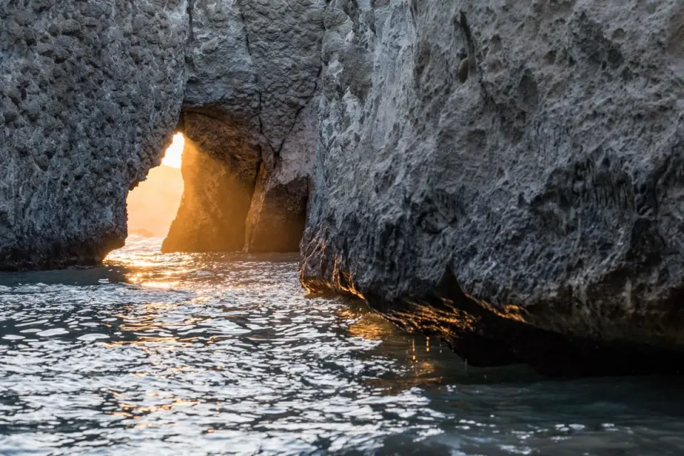 Golden sunset light shines through a rock arch, reflecting on water in Greece. - Photo by…